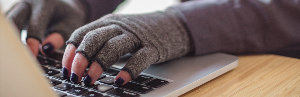 A disabled person types on a laptop while wearing compression gloves. They are lightskin and have black nail polish. The hands and keyboard are the focal point.